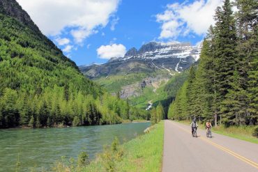 Two cyclists riding in Glacier National Park with mountains, trees, and a river nearby on a cycling holiday