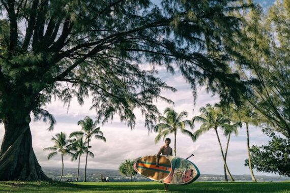 A man holds his inflatable SUP beneath palm trees near the shore