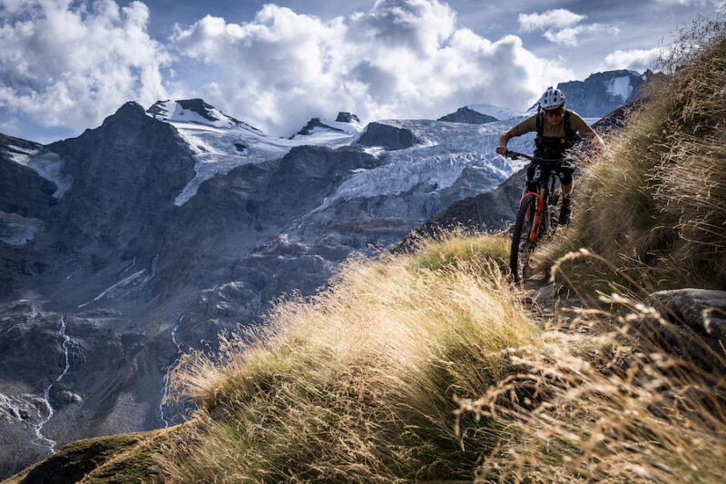 A mountain biker sprints down a trail, hugging the side of a mountain at sunset.