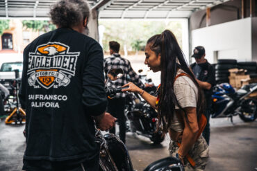 An EagleRider motorcycle expert shows a new female rider how to operate a motorcycle in a large garage.