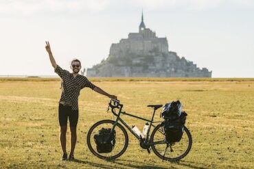 A young man standing next to his touring bicycle in an open field with a castle in the distance.