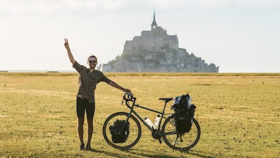 A young man standing next to his touring bicycle in an open field with a castle in the distance.