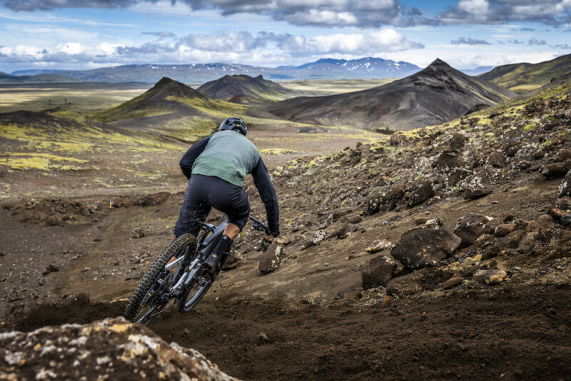 A mountain biker shreds a loose-dirt trail along a rock garden with mountain peaks in the distance in Iceland.