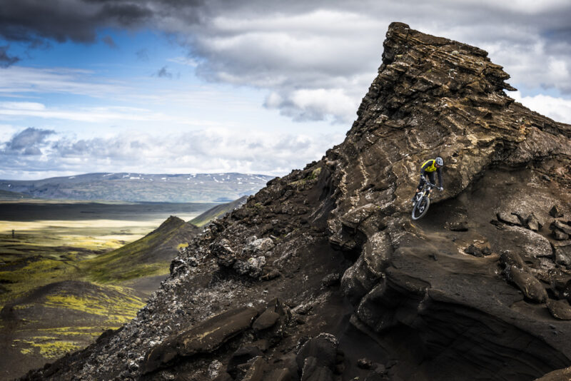 A mountain biker pedals along a rocky ledge with mountain peaks in the distance in Iceland.
