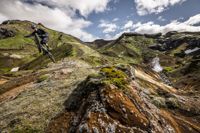 A mountain biker launches above a small, rocky hilltop in Iceland.