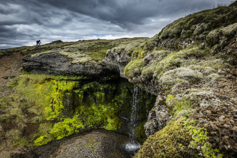 A mountain biker pedals along a mossy, rocky ledge with a waterfall below in Iceland.