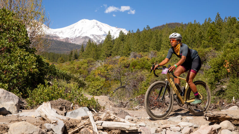  A female cyclist rides her gravel bike on a rocky trail with Mt. Shasta in the background