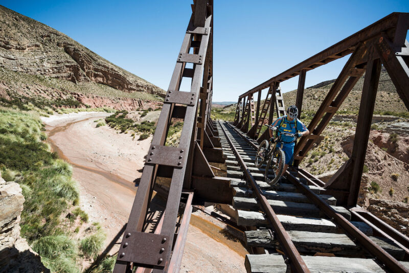 A mountain biker carrying his bike over a bridge with railroad tracks.