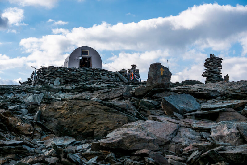 A small tin shed outpost atop a rocky cliff with two mountain bikers inside.