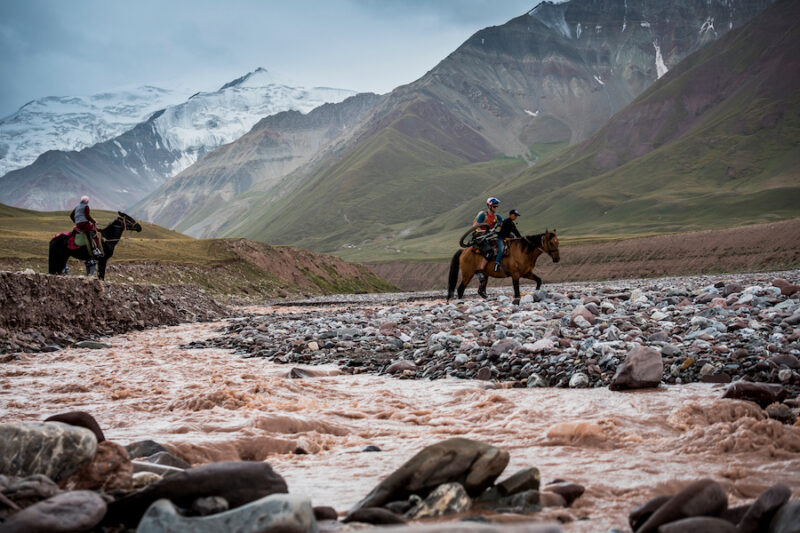 A mountain biker carries his bike on horseback through a rocky river crossing.