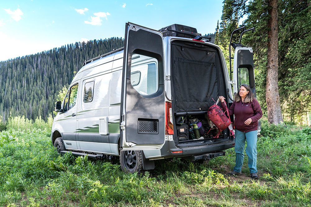 An older female standing at the rear of a Sprinter camper van with her outdoor gear.