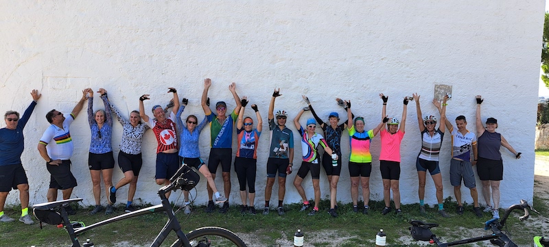 A large group of cyclists is standing against a big white wall with their hands in the air and smiling for the camera.
