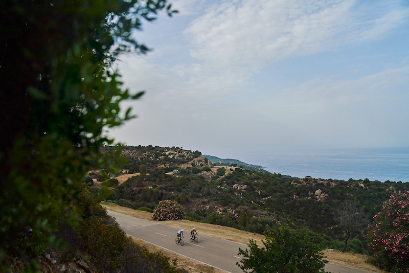 Two cyclists riding road bikes on an empty mountain road in Sicily with the coast in the background.