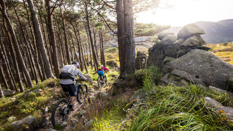 Two mountain bikers riding fast singletrack between trees and rocks in Scotland
