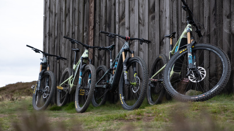 Four E-mountain bikes in a row in front of a rustic wood shed.