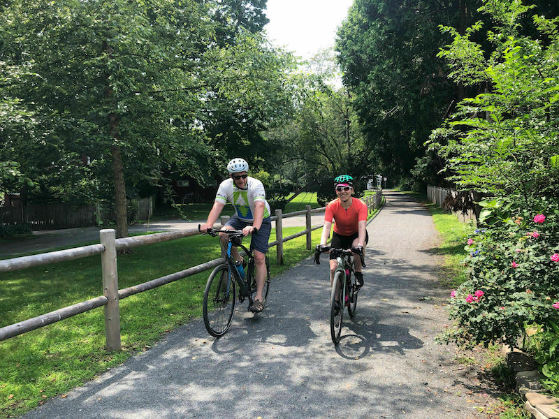 A male and female cyclist ride next to each other on a paved bike trail with a wood post fence on one side and trees in the background.