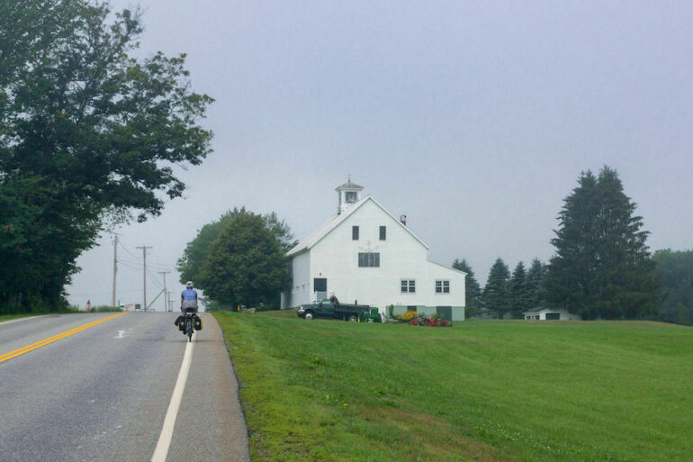 A lone cyclist riding on the shoulder of a two-lane highway in New England.