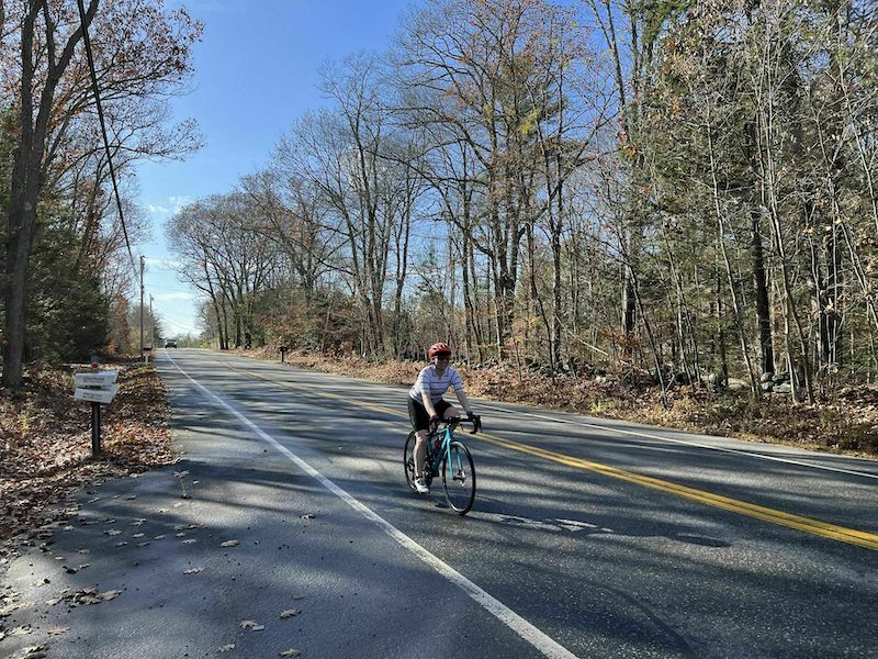 A female road cyclist riding on an empty Portland, Maine road with fall trees on both sides of her.
