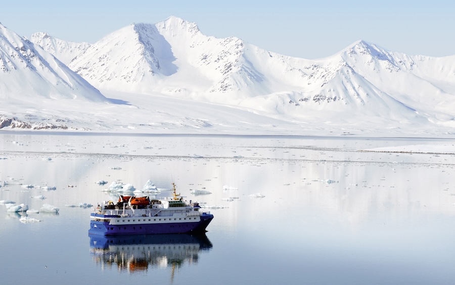 An expedition cruising ship idles on a glassy Arctic Ocean with snow-covered munitions in the distance.