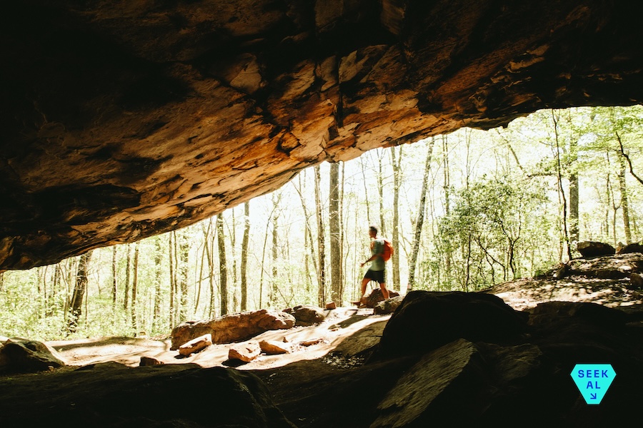 A solo hiker passes a massive cave opening on a rocky trail in Alabama's wilderness.